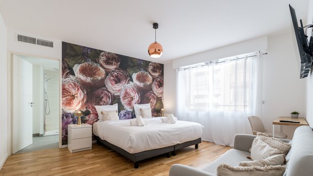 A bedroom with a large floral mural, a bed with white linens, a grey sofa, and a wooden floor.