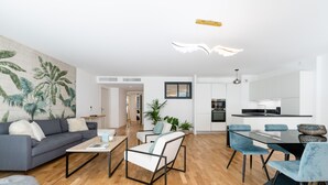A modern living room with a grey sofa, white armchairs, and a wooden coffee table. A kitchen with white cabinets and a dining area with blue chairs are visible in the background.