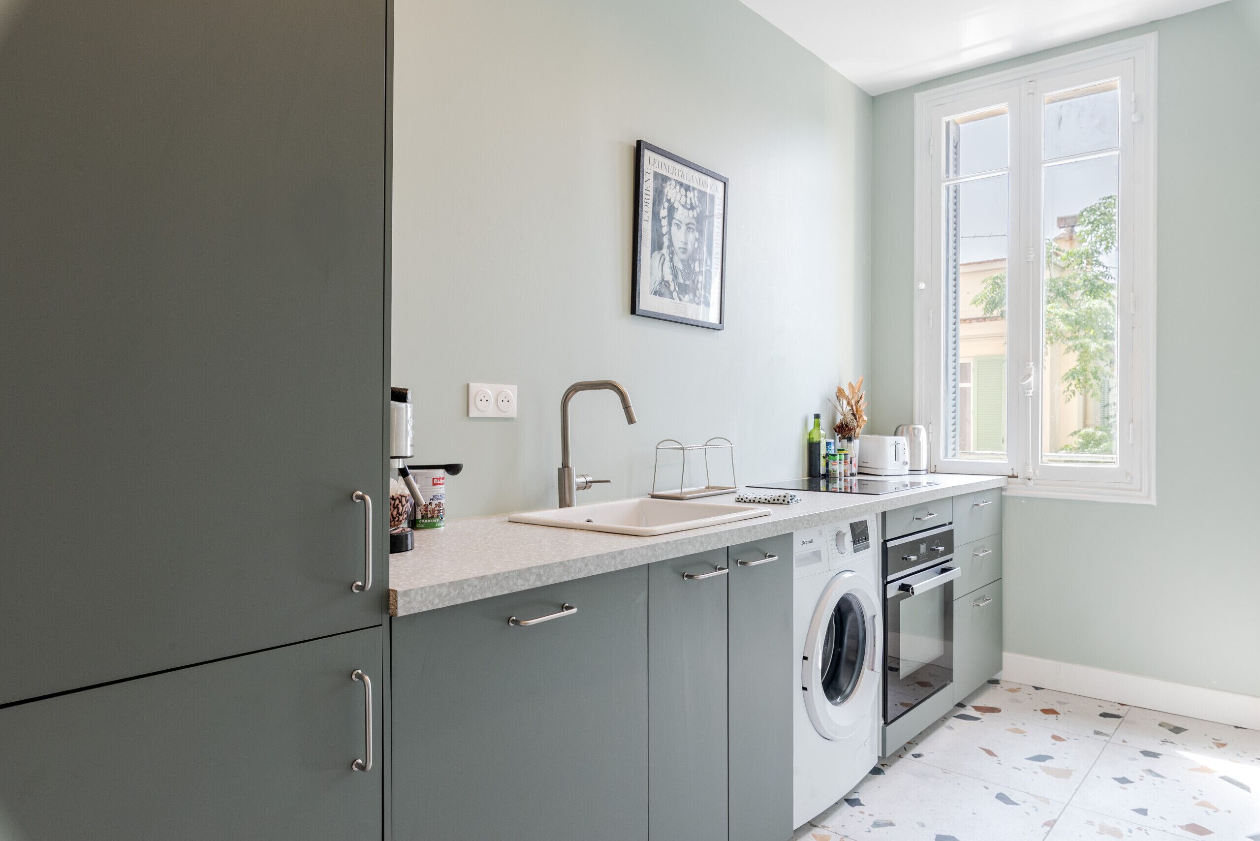 A modern kitchen with grey cabinets, a built-in washing machine, a sink, and a window with a view of greenery outside.