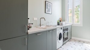 A modern kitchen with grey cabinets, a built-in washing machine, a sink, and a window with a view of greenery outside. - Angie By Terry's Homes (Cannes)