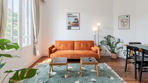 A living room with a sofa, two side tables, a coffee table, a dining table with chairs, a potted plant, and artwork on the walls. - Angie By Terry's Homes (Cannes)