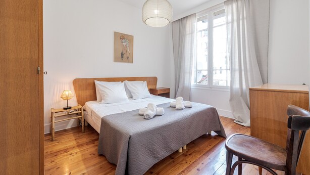 A bedroom with a wooden bed, a bedside table with a lamp, a chair, a desk, and a window with curtains. - Angie By Terry's Homes (Cannes)