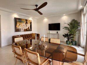 A modern living room with a glass dining table, wicker chairs, a brown sofa, a ceiling fan, a flat-screen TV, and a large painting.
