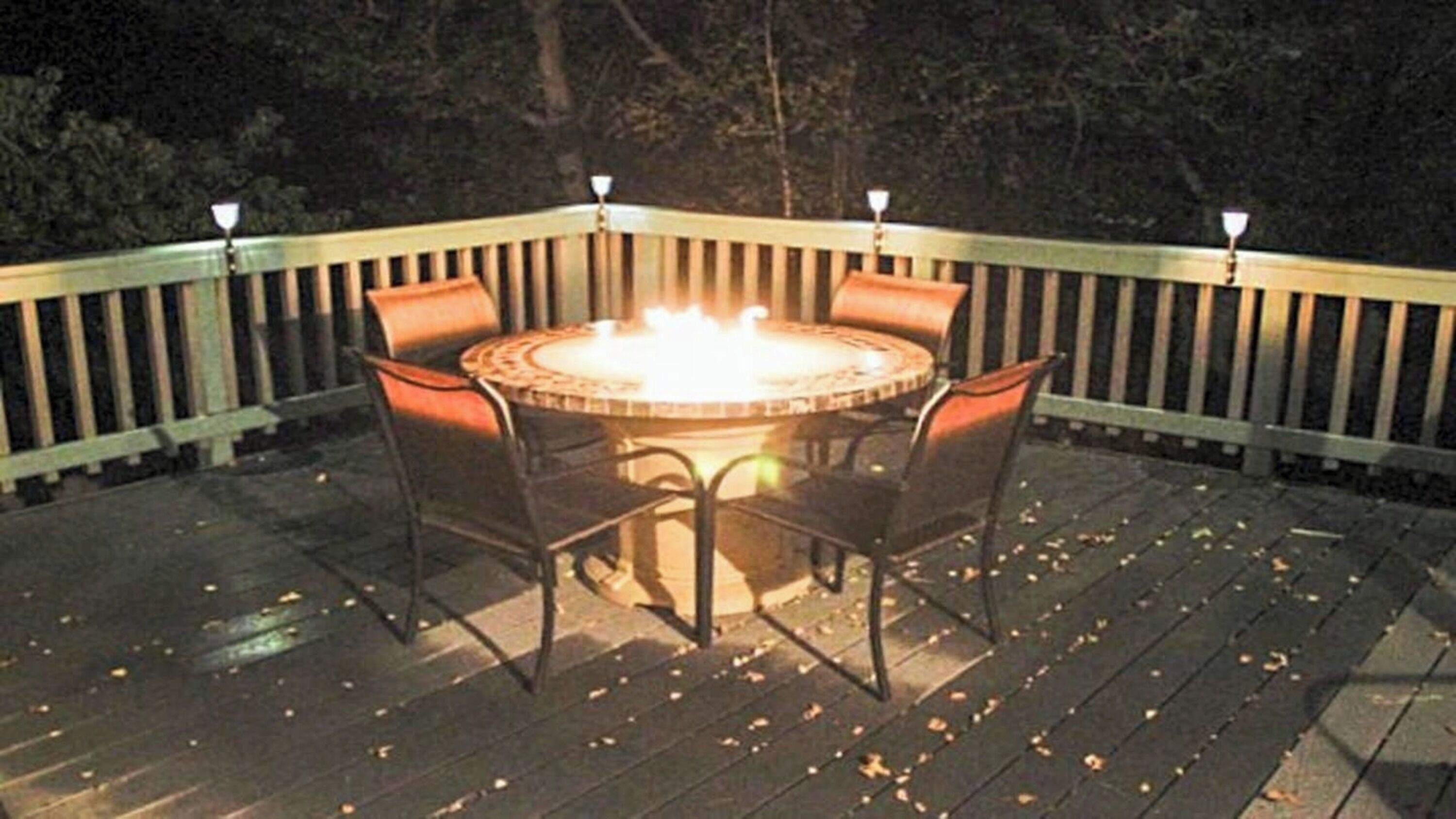 Et rundt bord med stole på en træterrasse, omgivet af gelænder med lys.