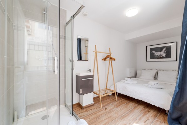 A modern hotel room with a glass shower enclosure, a wooden hanger, a bed with white linens, and a framed artwork on the wall.