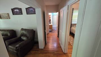 Un couloir au plancher de bois mène à une chambre à coucher qui contient un lit et une fenêtre. Deux fauteuils en cuir se trouvent sur le côté gauche, et une porte sur le côté droit.