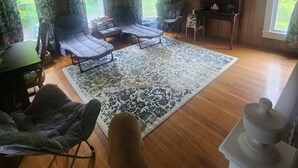 A spacious living room with a ceiling fan, large windows with floral curtains, a wooden floor, and a dog in the foreground.