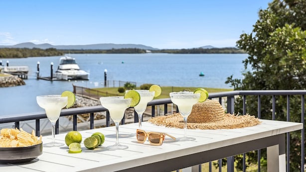 Un balcon donnant sur un lac, trois margaritas garnies de rondelles de citron vert, un bol de chips et une paire de lunettes de soleil posées sur une table.