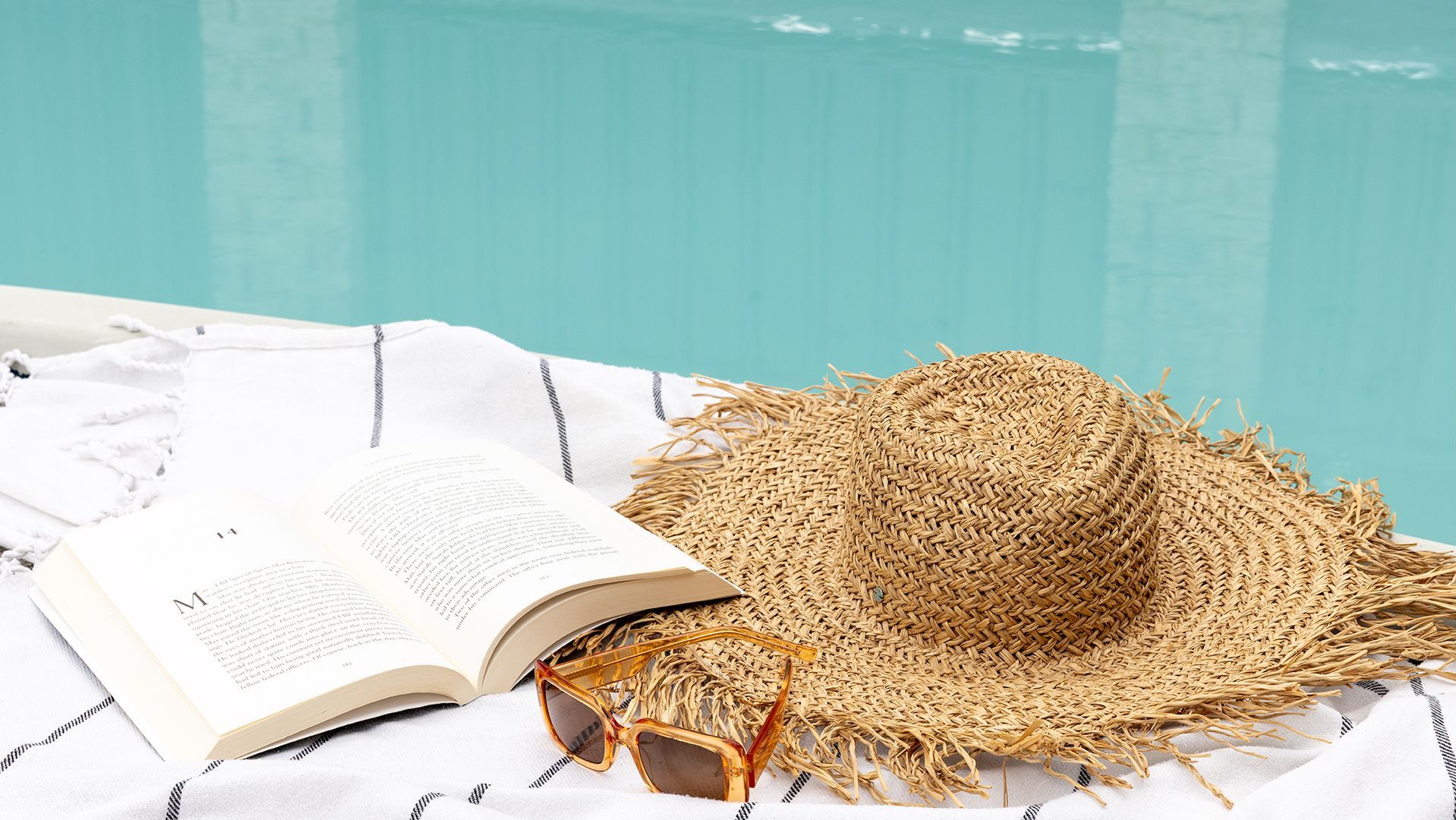 An open book, a straw hat, and sunglasses on a white towel by a pool.