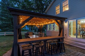 Une pergola en bois installée à l’extérieur, agrémentée de guirlandes lumineuses, avec une table en bois et des tabourets de bar.