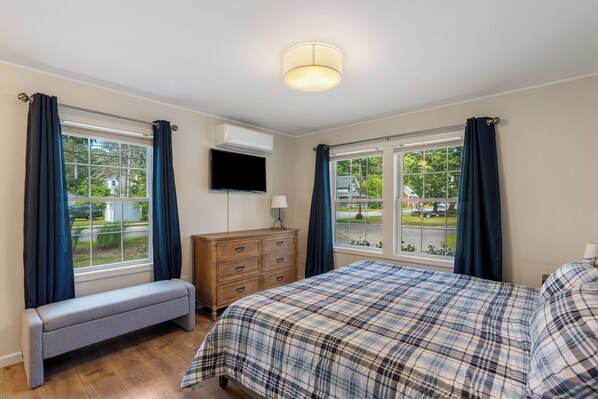 A bedroom with a bed, bench, dresser, TV, and large windows with curtains.