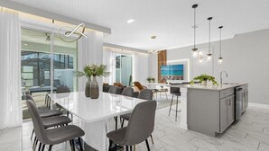 A modern dining area with a white table and grey chairs, a kitchen with grey cabinets, and pendant lights.