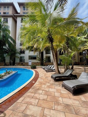 A pool area with lounge chairs, palm trees, and a building with balconies. - Villagio (Accra)