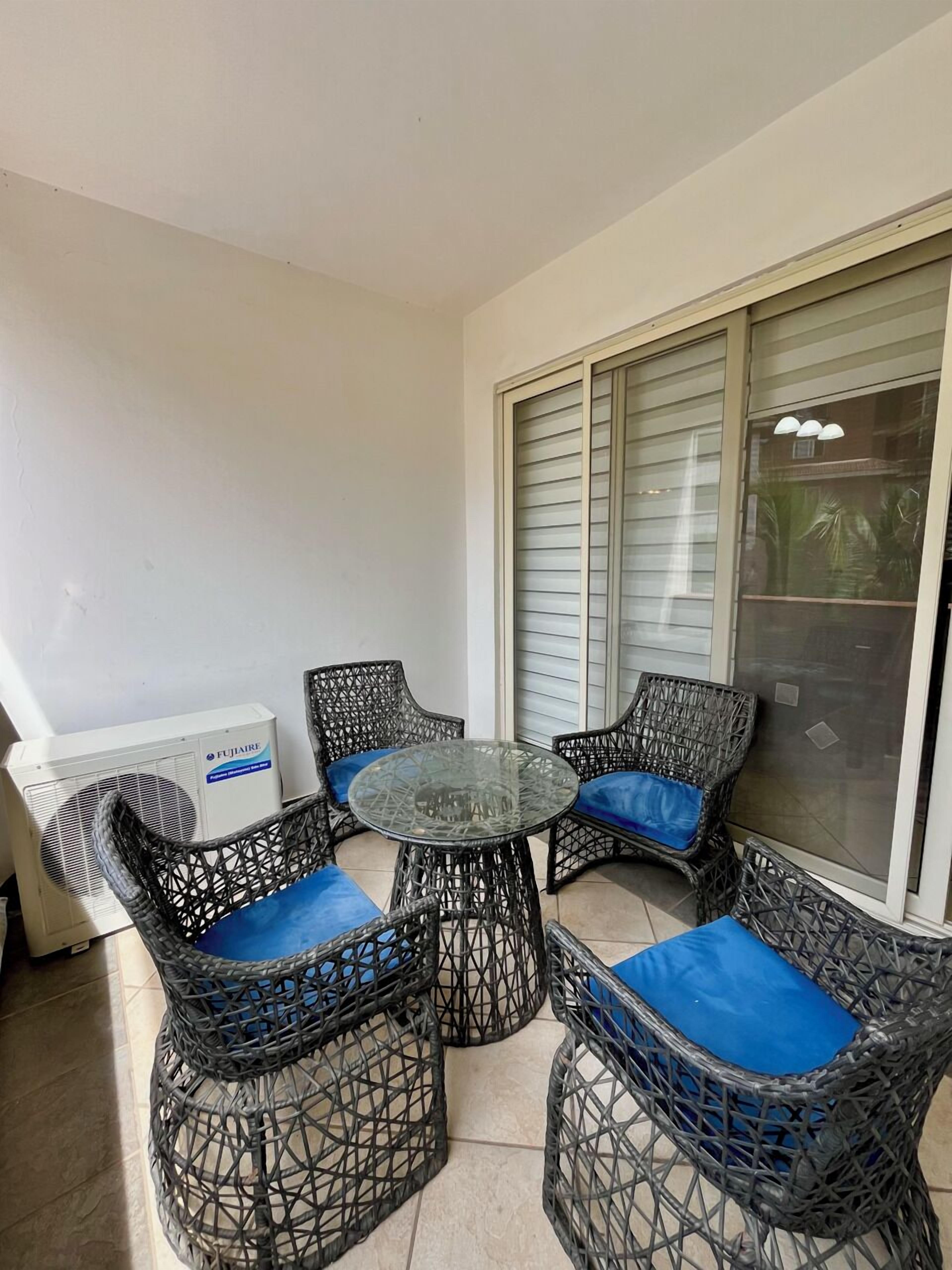 A balcony with wicker furniture, a glass table, and a white air conditioning unit.