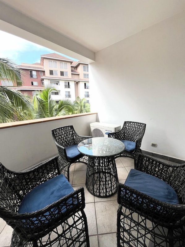 A balcony with wicker chairs and a glass table, overlooking a residential area with buildings and palm trees. - Villagio (Accra)