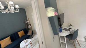 A modern living room with a dark blue sofa, a white coffee table, and a chandelier. There is a desk with a computer and a chair, and a small plant on the desk. - Provencal style suite (second floor) (Licata)