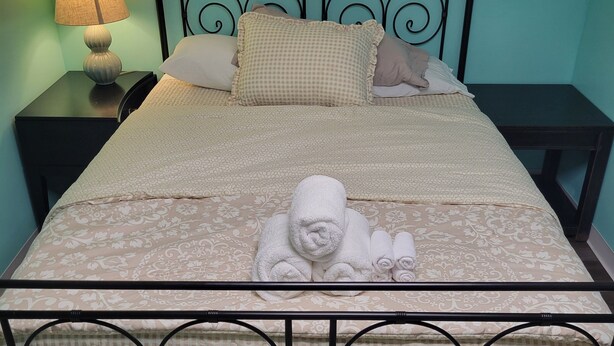 Una cama bien hecha con una colcha estampada, almohadas y una toalla enrollada. Hay un aire acondicionado de pared sobre la cama y una mesita de noche con una lámpara.