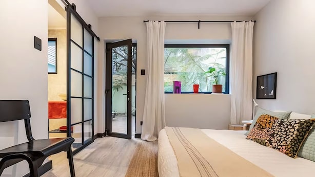 A bedroom with a bed, a chair, a window with curtains, a desk, and a potted plant.
