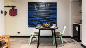 A dining area with a table set for two, a large abstract painting on the wall, and a kitchen area to the right.