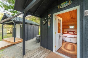 A wooden cabin with a porch, a small outdoor seating area, and a visible bedroom with a bed and storage.