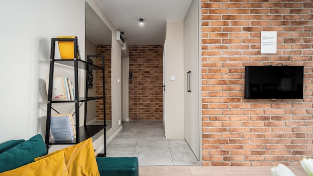 Un salón con pared de ladrillo, un televisor de pantalla plana montado en la pared, una estantería y un sofá verde con cojines amarillos.