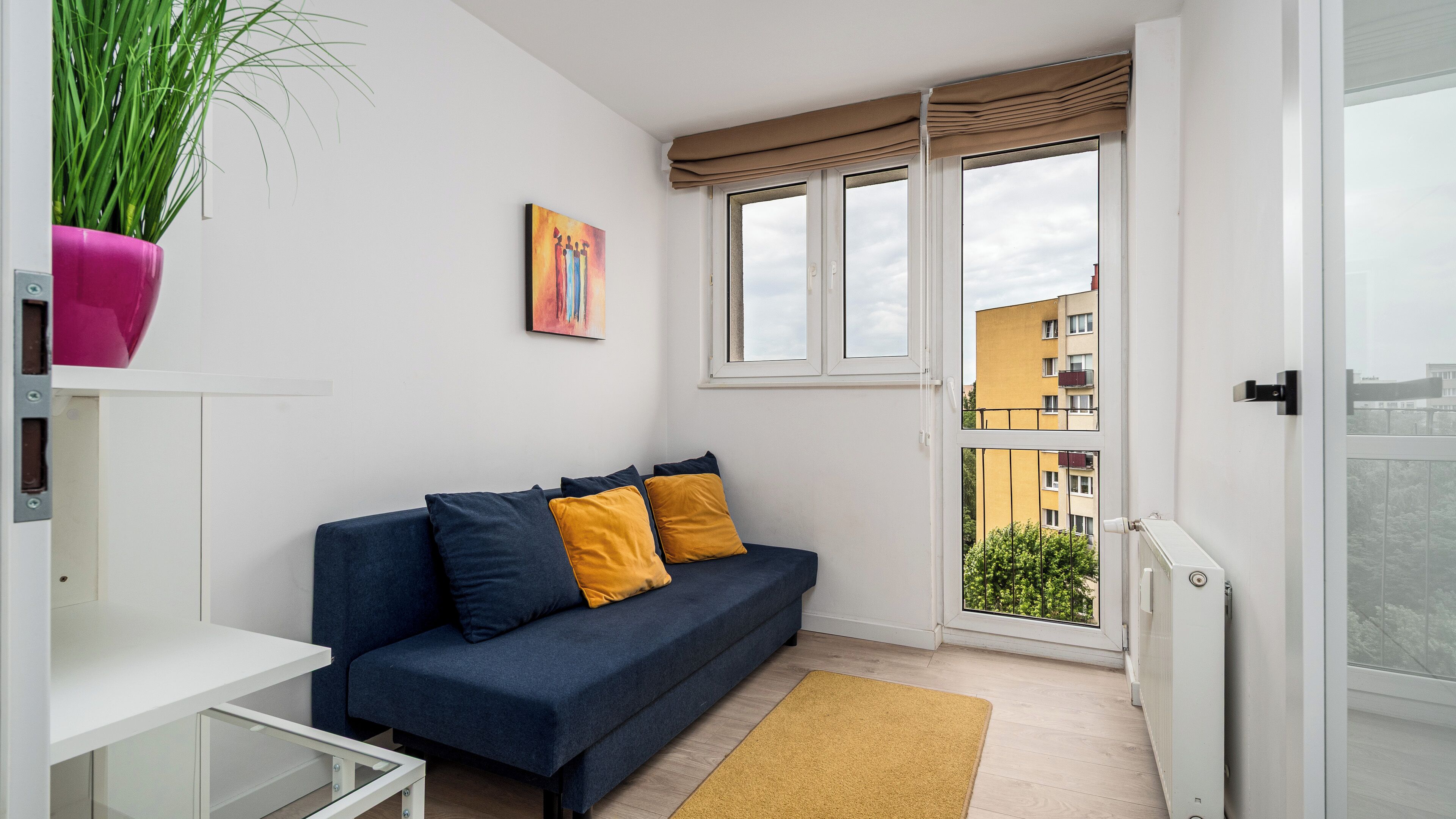 A small, modern apartment with a dark blue sofa, yellow cushions, a white shelf, a potted plant, and a painting on the wall.