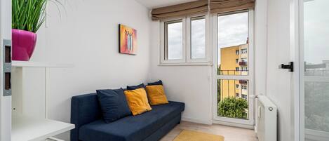 A small, modern apartment with a dark blue sofa, yellow cushions, a white shelf, a potted plant, and a painting on the wall.