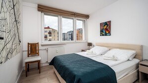 Un dormitorio con cama, una silla, una ventana con vista a edificios y un cuadro en la pared.