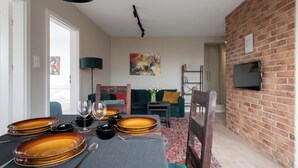 Un área de comedor con una mesa lista para dos personas, un sofá, un televisor montado en una pared de ladrillo y una estantería.