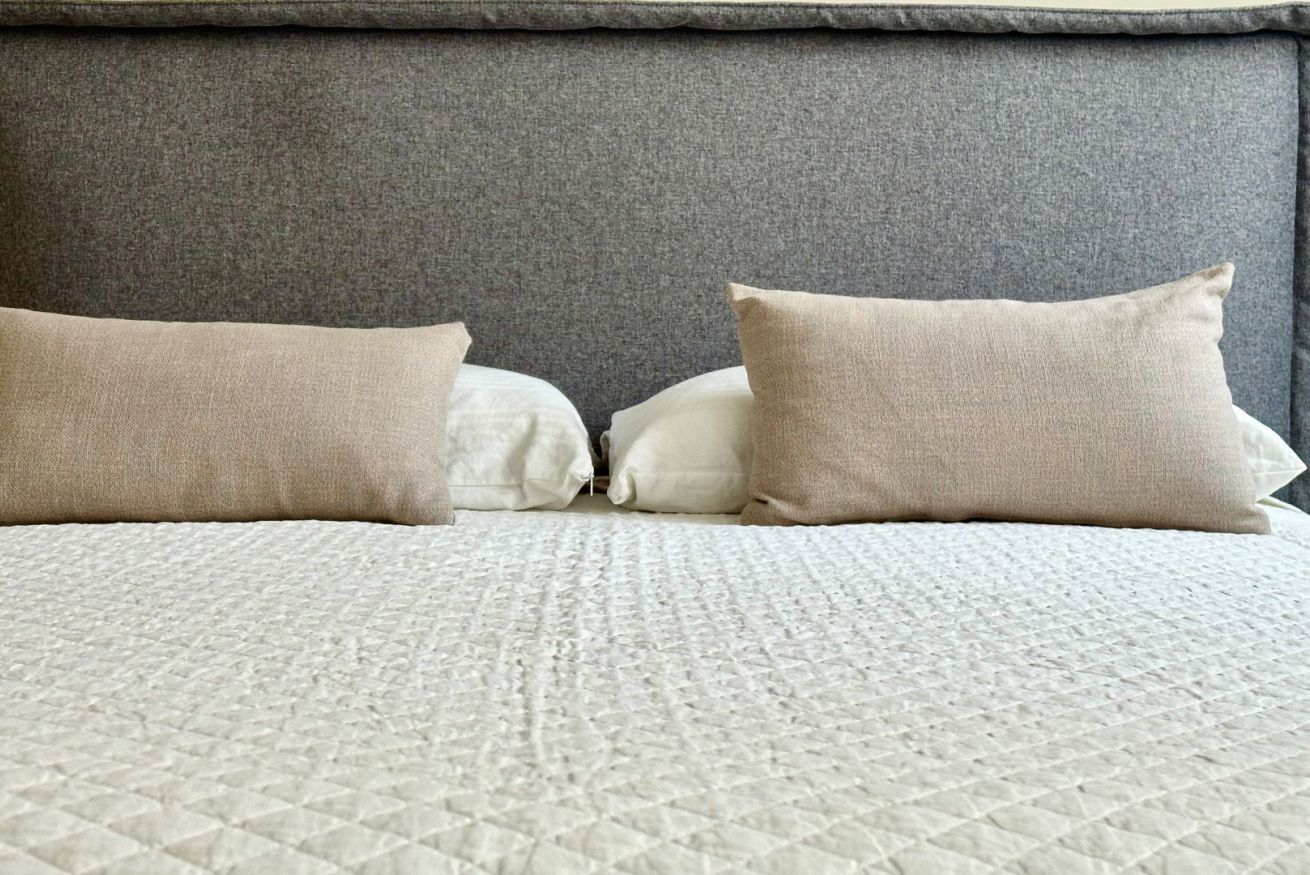 A bed with a quilted bedspread and two pillows, one white and one beige, against a grey headboard.