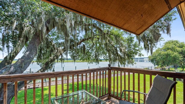 A wooden deck with a glass table and chairs, overlooking a lake with trees and a distant building.
