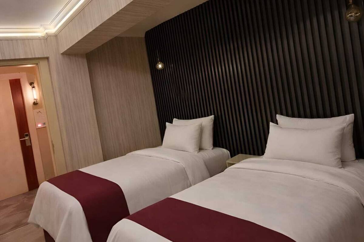 Habitación de hotel con dos camas, cabecera de madera oscura y pared de color claro.