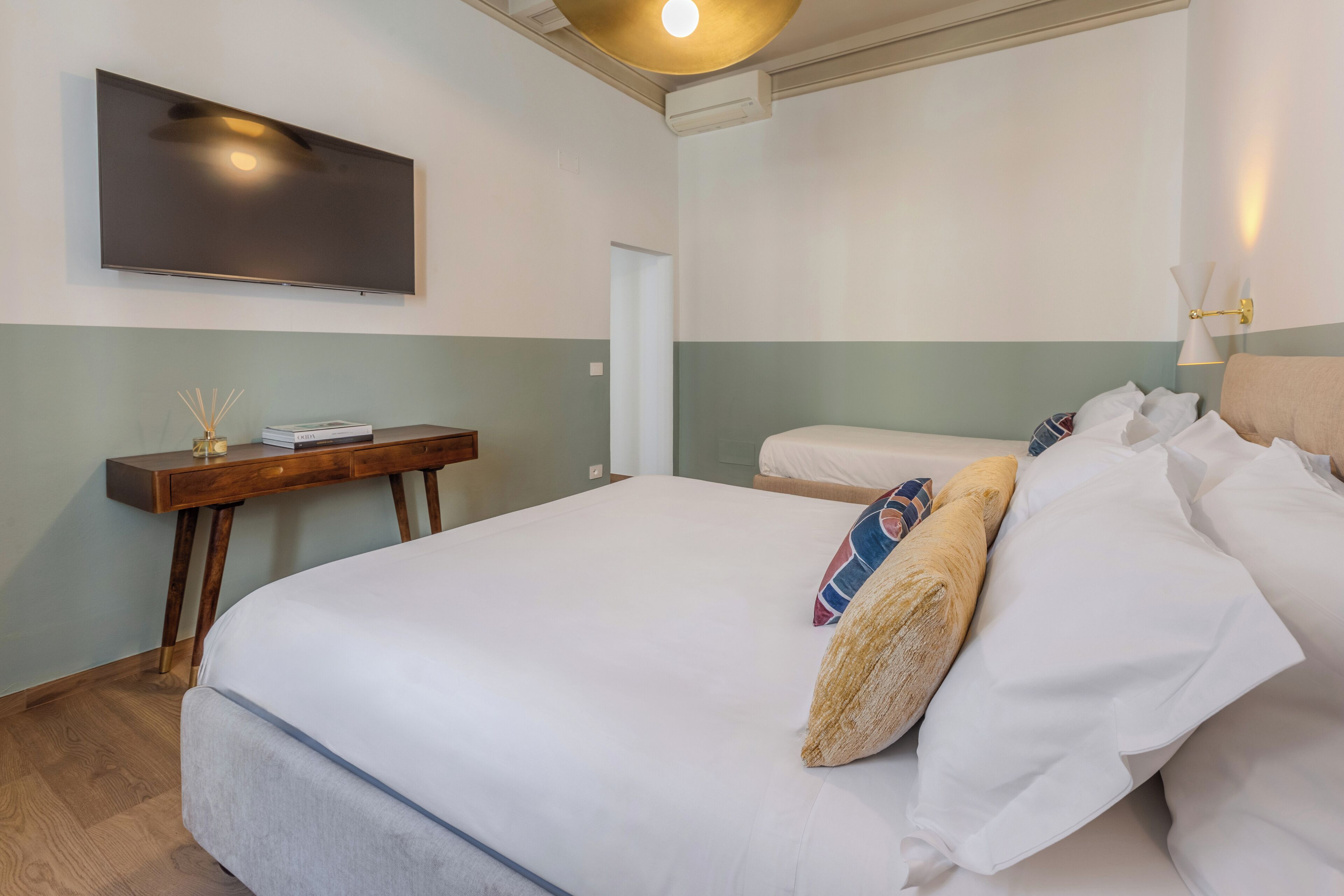 A hotel room with a large bed, a bedside table with a book and a vase, a flat-screen TV mounted on the wall, and a wall-mounted lamp.