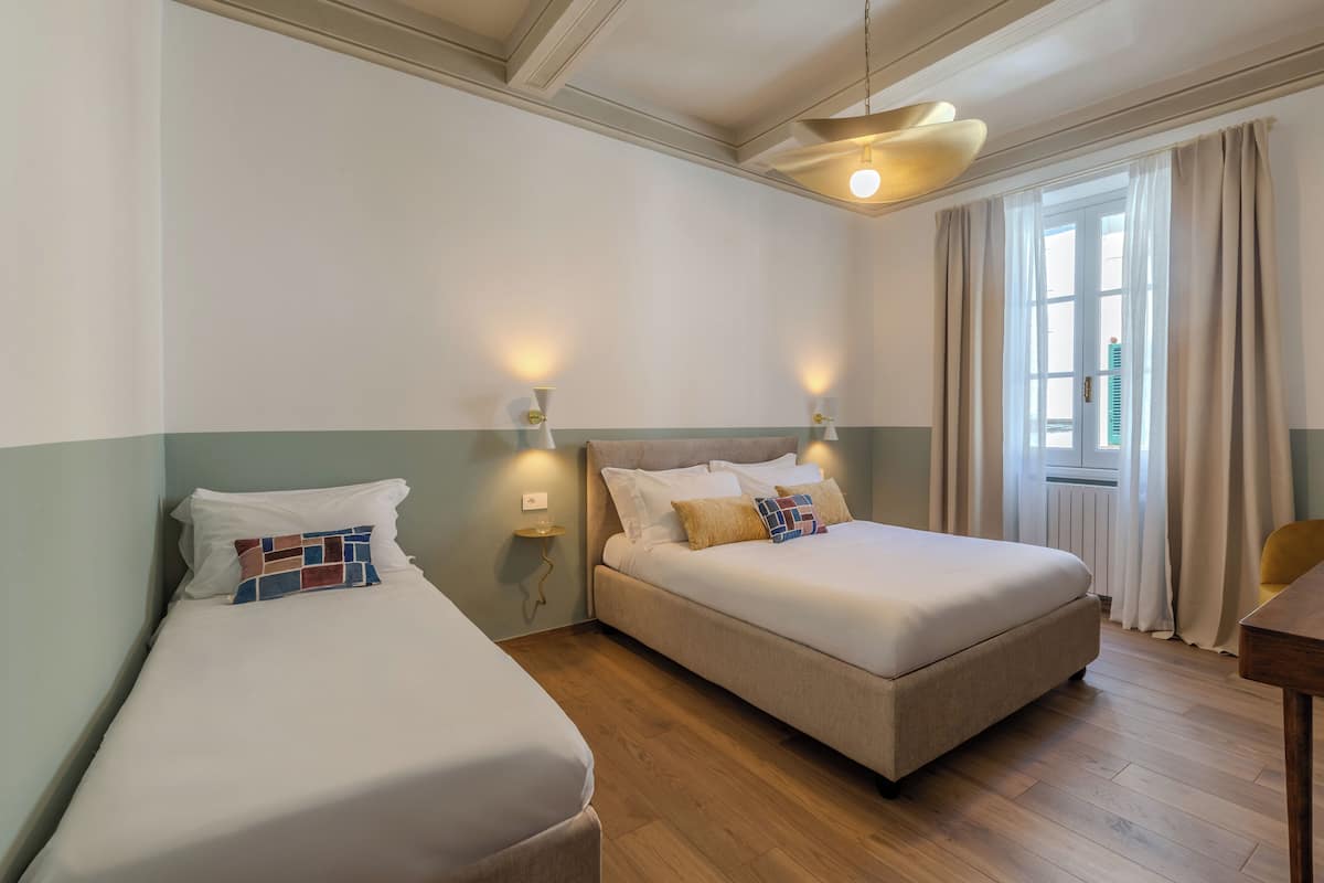 A hotel room with two beds, a wooden floor, a window with curtains, and a wall-mounted light fixture.