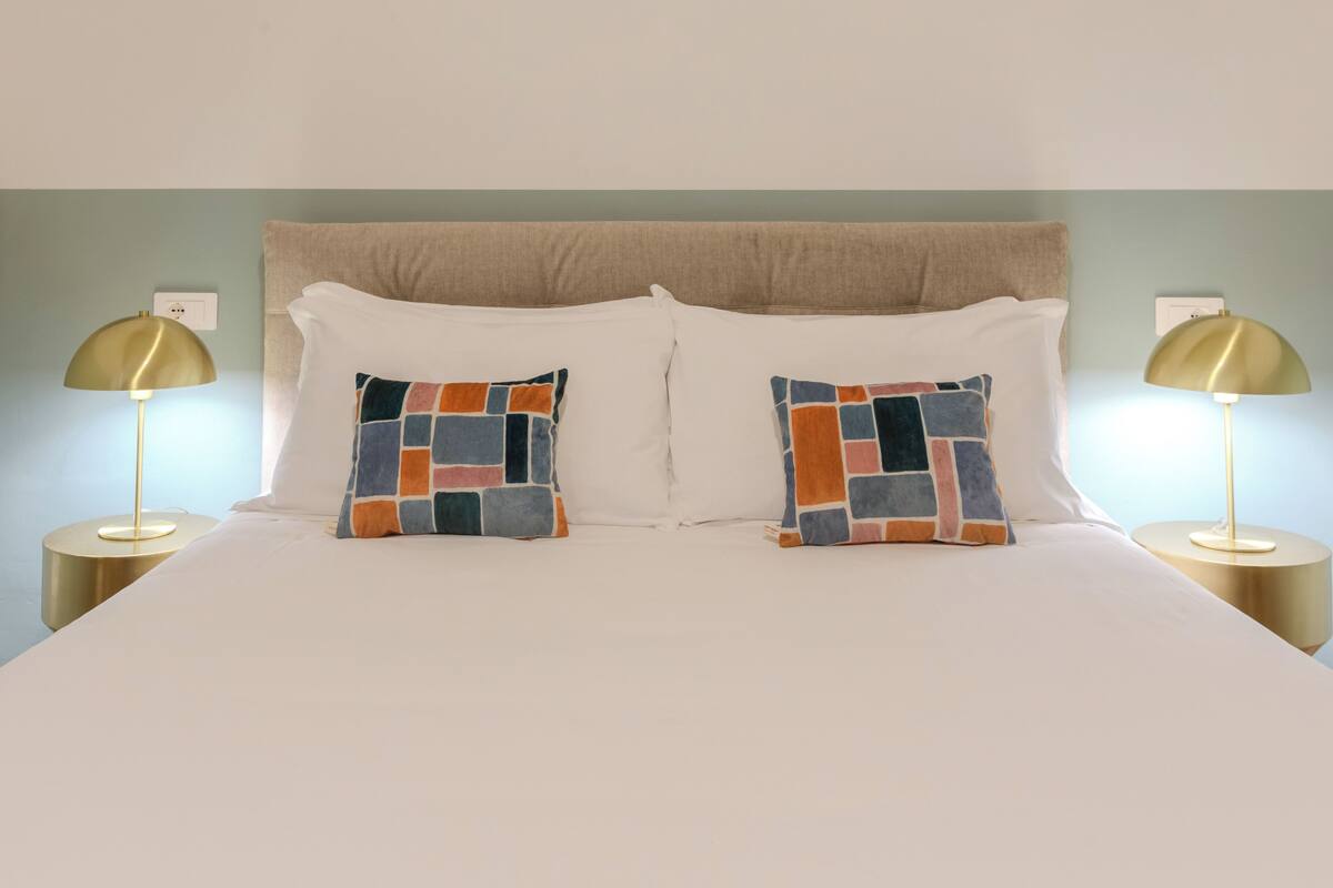 A neatly made bed with white linens and two decorative pillows, flanked by two gold lamps on matching side tables.