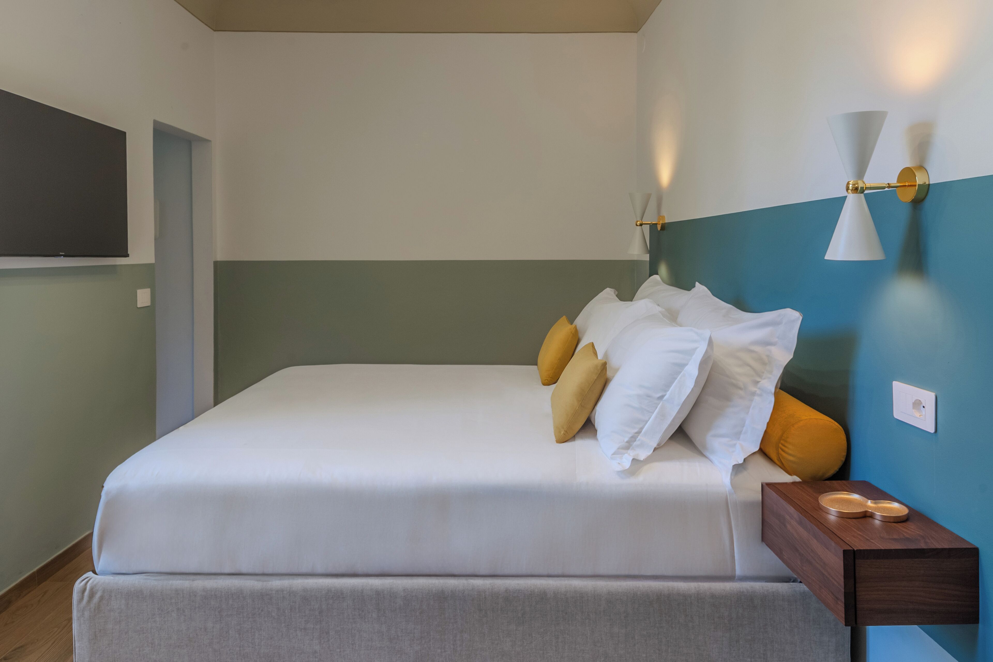 A neatly made bed with white and yellow pillows, a wooden nightstand with a bowl, and a wall-mounted light fixture.