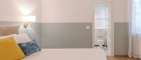 A hotel room with a bed, a wall-mounted lamp, a towel, and a view of the bathroom.