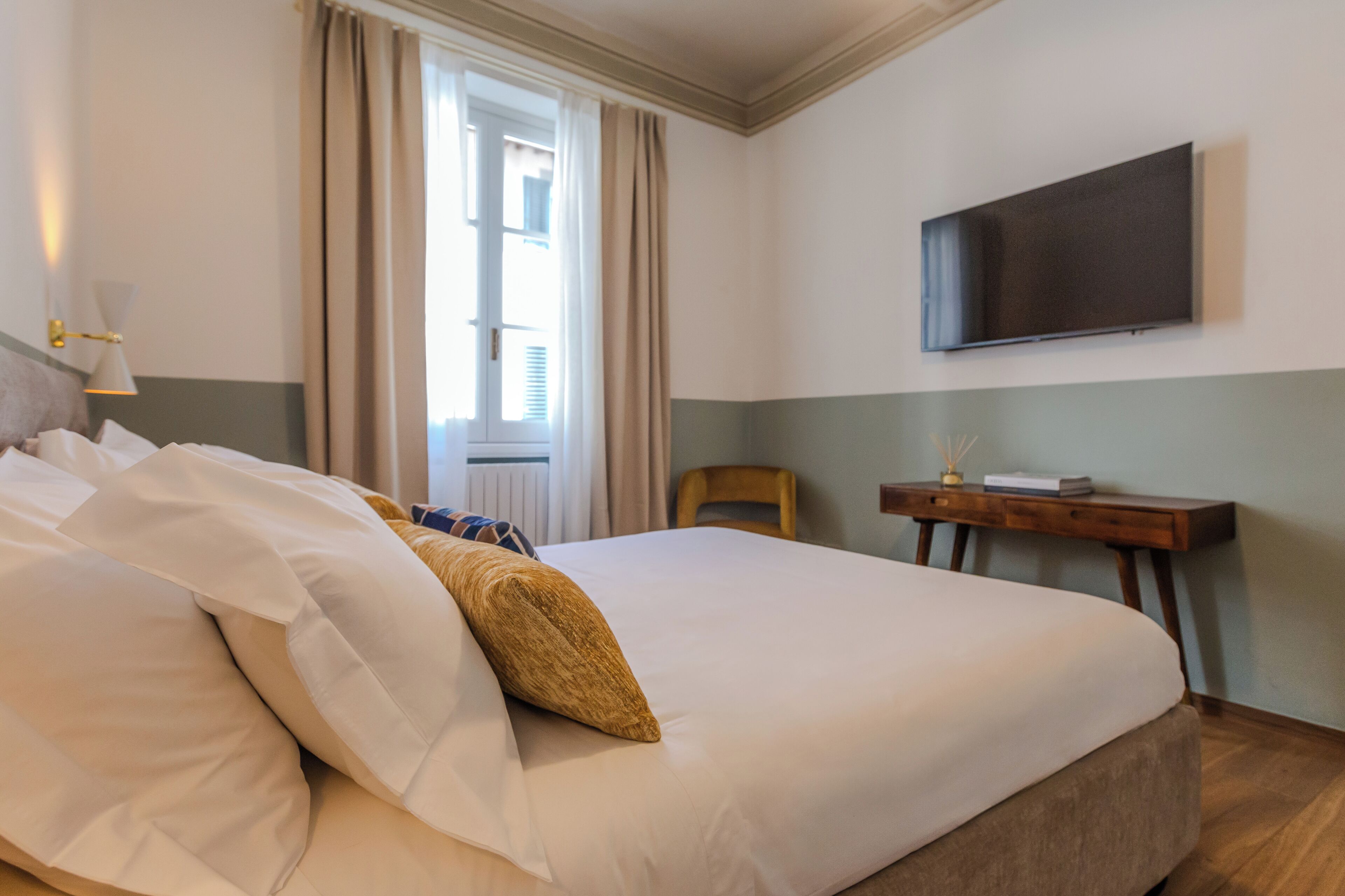 A neatly made bed with white and beige bedding, a bedside table with a book and a lamp, and a flat-screen TV mounted on the wall.