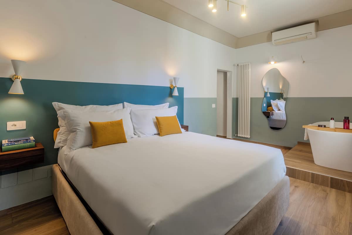 A modern hotel room with a large bed, bedside tables, a built-in bathtub, and a mirror on the wall.
