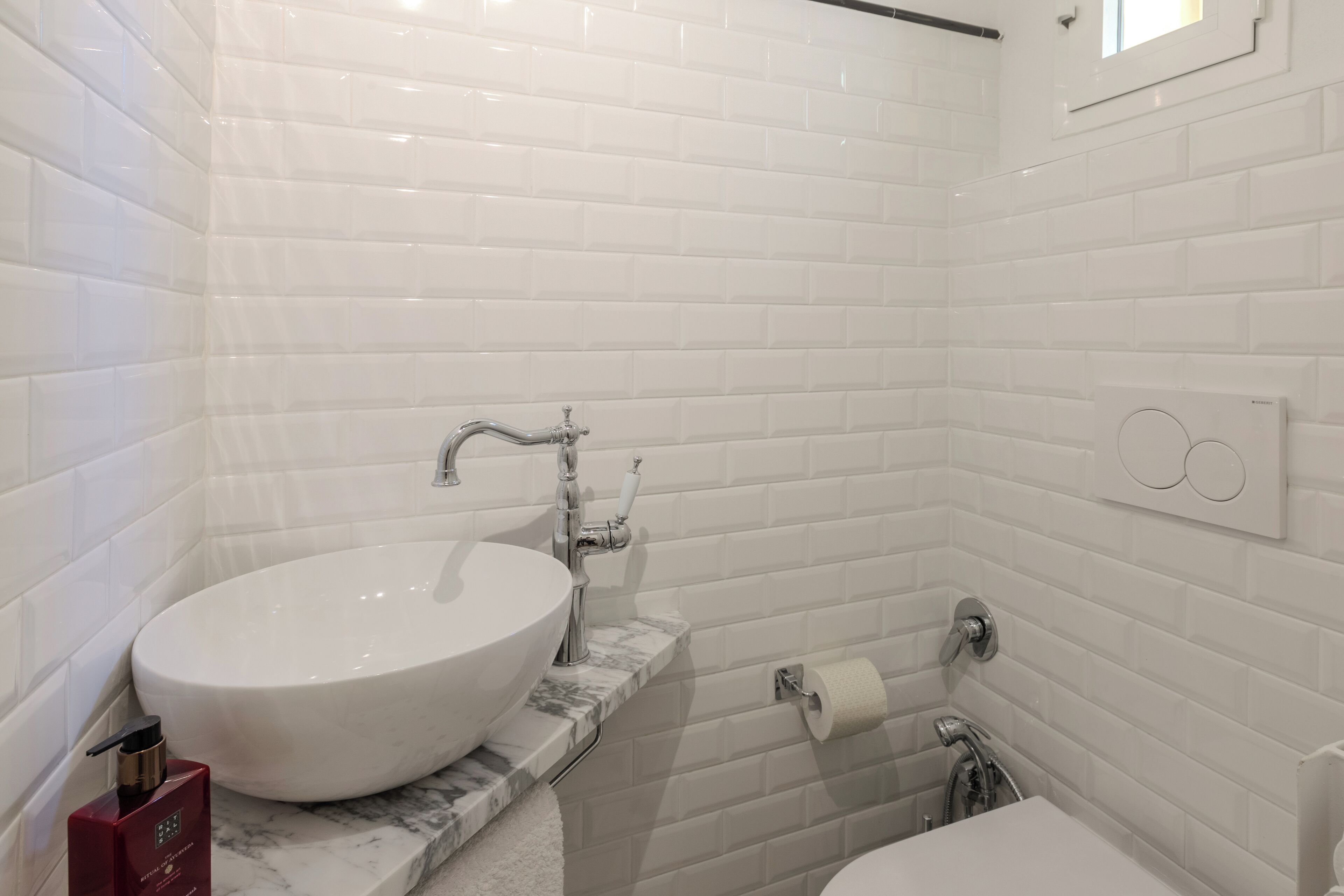 A bathroom with white subway tiles, a round sink, a marble countertop, a towel rack, and a toilet paper holder.