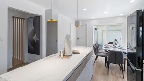 Una cocina moderna con encimera de mármol, iluminación colgante y comedor separado por un panel de vidrio.