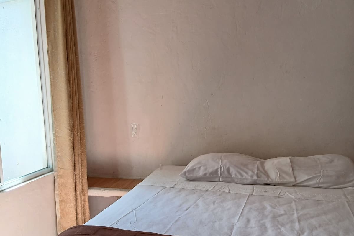 A bedroom with a bed, a window with curtains, a wall-mounted air conditioner, and a power outlet.