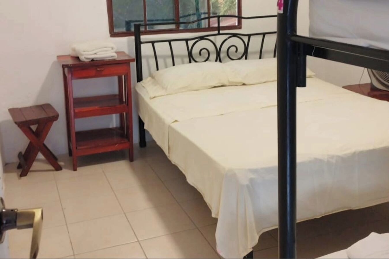 A single bed with a metal frame and a white mattress, a wooden nightstand with a folded white towel, and a small wooden stool.