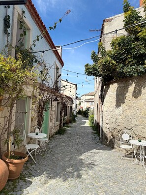 A narrow cobblestone street flanked by buildings with green shutters and white walls, featuring outdoor seating areas with metal chairs and tables. - Sadele Hacımemiş (Cesme)