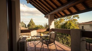 Eine überdachte Terrasse im Freien mit Tisch und Stühlen, ein kleiner runder Tisch mit einer Schale und Bäumen sowie Gebäuden im Hintergrund.