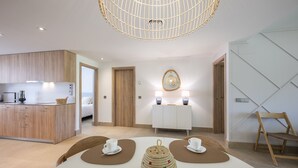 Ein modernes Hotelzimmer mit einer Küchenzeile, einer Essecke mit zwei weißen Stühlen und einem gewebten Hocker, einem Holzstuhl und einer außergewöhnlichen Deckenleuchte.