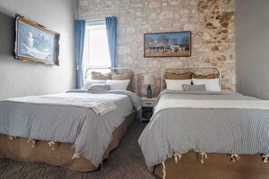 A room with two beds, a stone wall, a window with curtains, and two framed pictures. - The Bank-Luxury Suite w/Deck Overlooking Main St! (Fredericksburg)