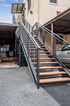 Un escalier métallique mène à l’entrée d’un immeuble, et une voiture est garée à proximité.