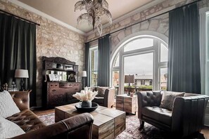 A spacious living room with a stone wall, leather furniture, and a large arched window offering a view of the city. - The Bank-Luxury Suite w/Deck Overlooking Main St! (Fredericksburg)