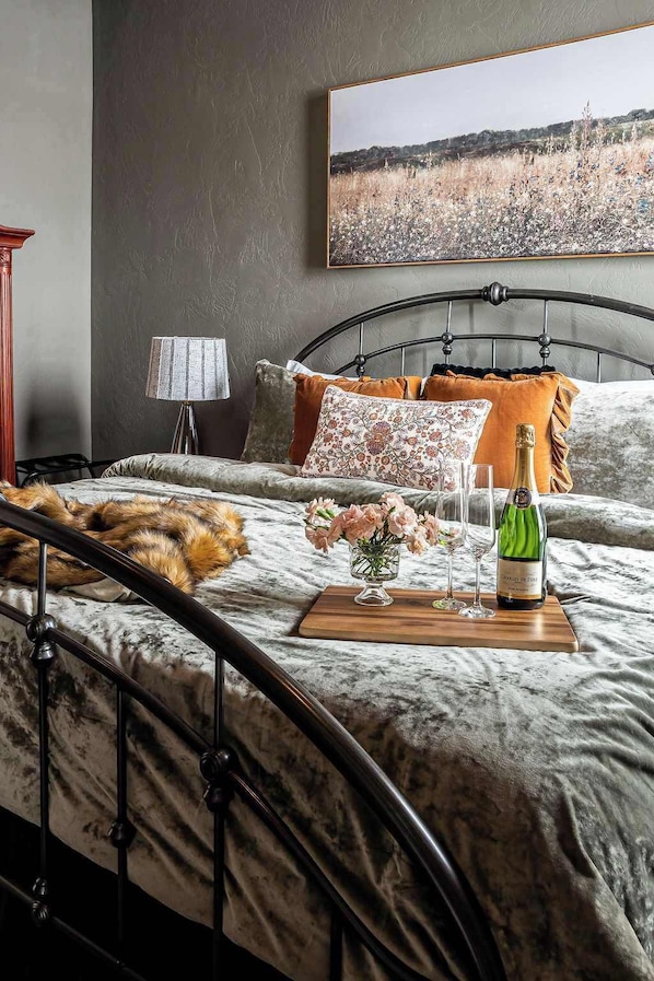 A bedroom with a bed, a nightstand, a lamp, a framed picture on the wall, and a bottle of champagne on the bed.